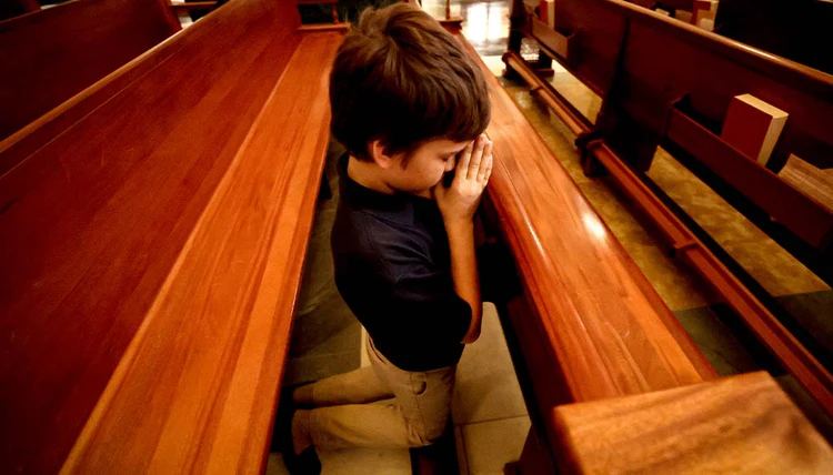 Young boy kneeling in prayer at a wooden church pew with his hands clasped, showing deep devotion and faith.